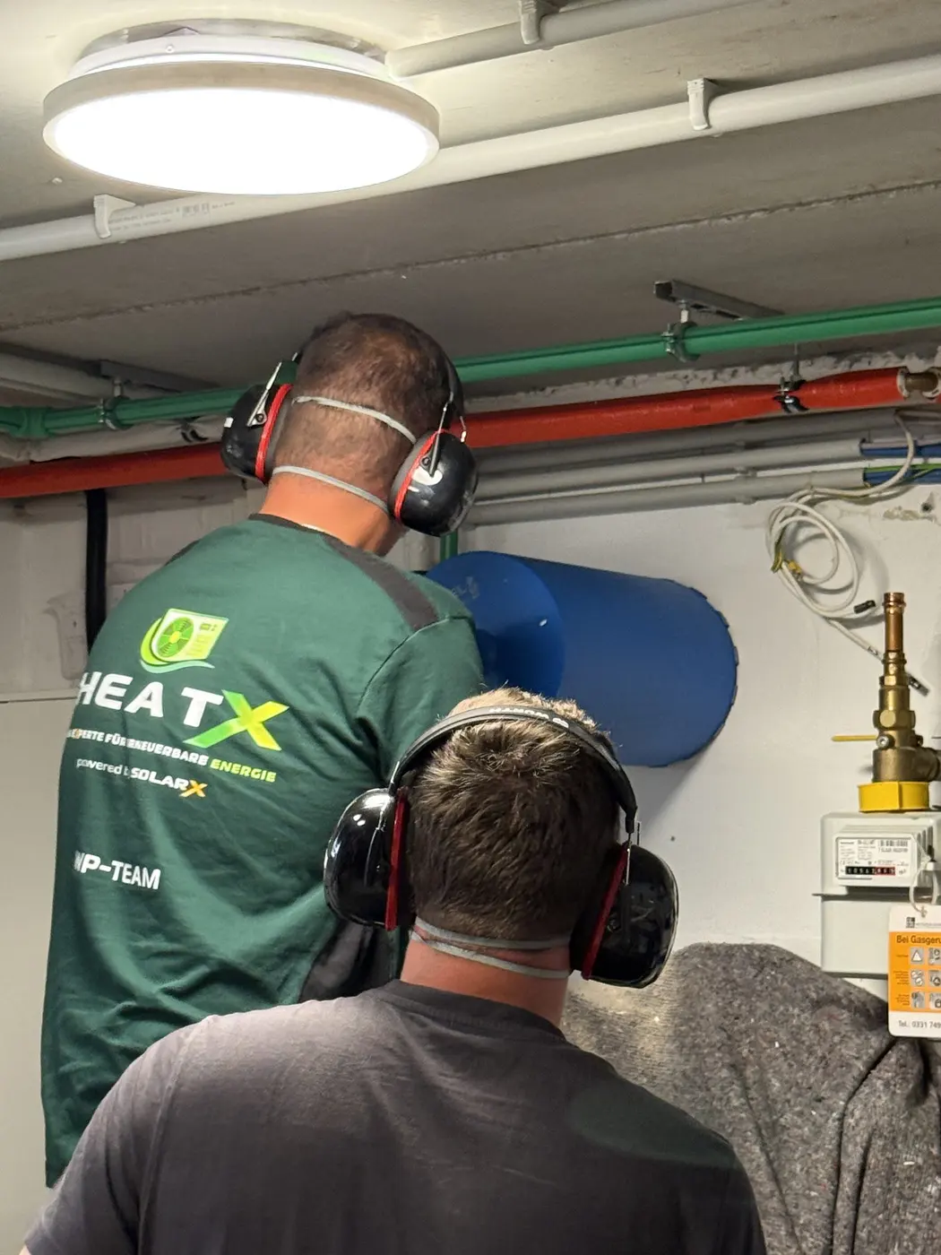 HeatX – Monteure bei der Kernbohrung für eine Wärmepumpe HeatX Monteure in Arbeitskleidung mit Gehörschutz bei der Kernbohrung für eine Wärmepumpe im Keller