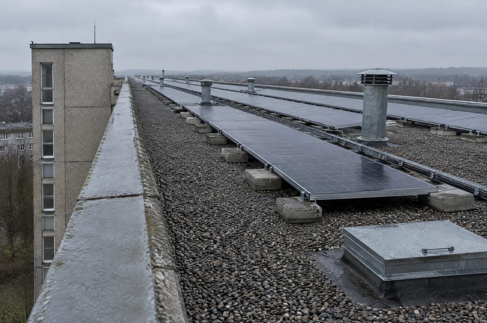 Ein großer Teil des Gebäudebestands entstand nach dem Zweiten Weltkrieg, darunter zahlreiche Plattenbauten. Bei diesen Gebäuden prüfen wir insbesondere Dachzustand, Statik und die geeignete Befestigung der Solarmodule. Nahe Sicht auf das Flachdach eines zehngeschossigen Plattenbaus in Cottbus: bekieste Bitumenbahnen, umlaufende Attika, Lüftungsrohre und ein Treppenhauskopf. Dunkle Solarmodule stehen paarweise in niedrigen Ost-West-Wannen, mit Betongewichten beschwert und ohne Durchdringung der Dachhaut.