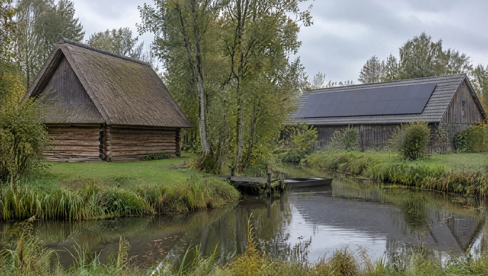 Im Süden von Dahme-Spreewald, um Straupitz, Schlepzig und Lübben, besteht der Bestand aus Blockhäusern, Vierseithöfen und Scheunen. Das Rohrdach ist dort steil gedeckt, damit das Wasser abläuft — weiche Bedachung, Brandschutz und die fehlende tragende Unterkonstruktion machen eine Belegung zur Ausnahme. Die nutzbare Fläche liegt deshalb meist auf dem Nebengebäude: Scheune, Stall oder Remise, hart gedeckt, groß und ungeteilt. Das Bild stellt genau diese Arbeitsteilung auf einem Hof nebeneinander. Breite Aufnahme einer Hofstelle im südlichen Dahme-Spreewald: links ein Blockbohlenhaus mit steilem Reetdach ohne Module, rechts eine Scheune mit flacherem Satteldach aus grauen Betondachsteinen, darauf eine geschlossene Modulfläche; dazwischen Erlen und ein stilles Fließ.