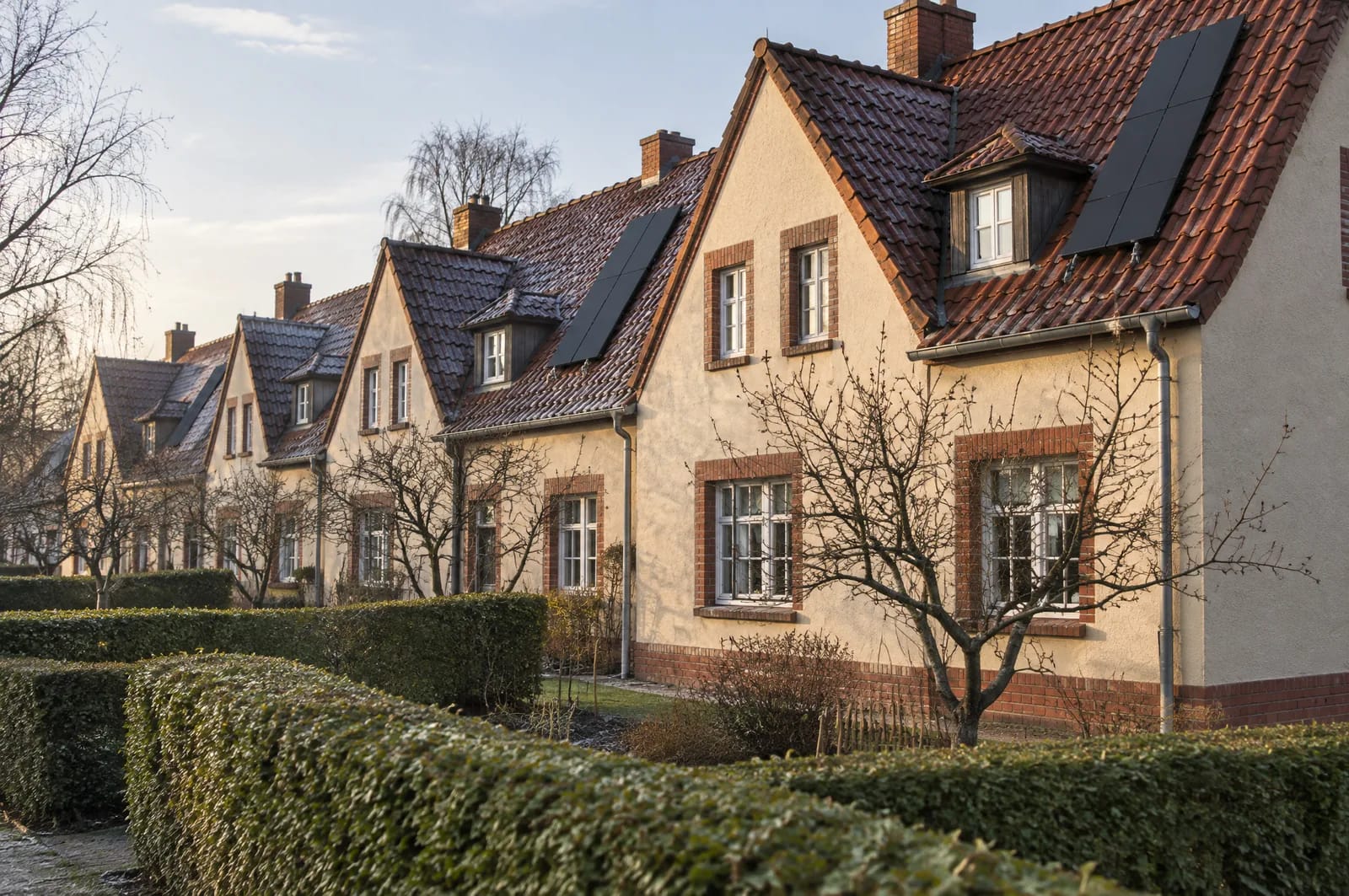 Als die Reichsbahn nach dem Ersten Weltkrieg ihre Direktion Osten nach Frankfurt verlegte, brauchte sie Wohnraum für mehr als 600 Familien; ab 1922 entstanden dafür eigene Siedlungen mit Reihen- und Mehrfamilienhäusern. Ihre Dächer sind das Gegenteil der Innenstadt: steil geneigt, mit Tonziegeln gedeckt, durch Schleppgauben, Schornsteine und Brandwände in kurze Abschnitte geteilt. Hier zählt jedes einzelne Modul, die Belegung folgt der nutzbaren Restfläche zwischen Gaube und First, und bei Ensembleschutz redet die untere Denkmalschutzbehörde mit. Nahansicht einer zweigeschossigen Reihenhauszeile im Stil der Eisenbahnersiedlungen der 1920er Jahre in Frankfurt (Oder): cremefarbener Putz, Ziegelsockel, steile Satteldächer mit roten Tonziegeln, kleine Schleppgauben und darauf ein geschlossenes Feld schwarzer Module zwischen Gaube und First.