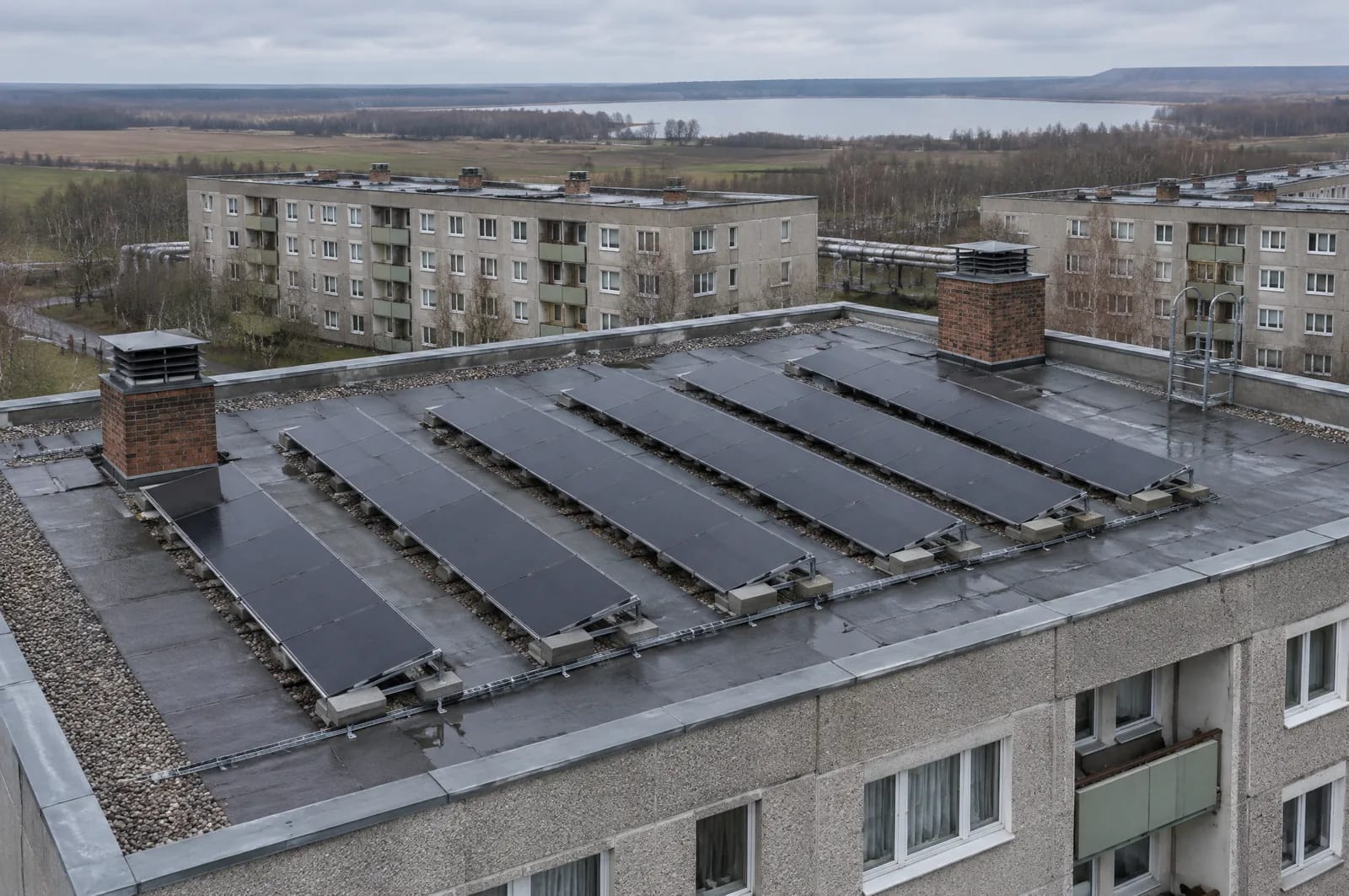 Zwischen 1946 und 1975 entstanden allein in Lübbenau rund 6.000 ferngeheizte Wohnungen in Plattenbauweise, in Senftenberg kamen zwischen 1980 und 1986 etwa 3.055 Wohnungen hinzu, dazu die Vetschauer Neustadt. Das Flachdach ist damit im Landkreis Oberspreewald-Lausitz kein Randfall, sondern ein eigener Bautyp. Es gibt die Ausrichtung frei, verlangt dafür eine Aussage zur Statik und zum Zustand der Bitumenbahn, bevor Ballastgestelle aufgesetzt werden. Aufgeständerte Reihen brauchen zudem Abstand gegen die gegenseitige Verschattung. Nahsicht auf das bitumengedeckte Flachdach eines fünfgeschossigen Plattenbaus im Landkreis Oberspreewald-Lausitz: sechs Reihen aufgeständerter schwarzer Module auf Ballastgestellen, Kiesstreifen an der Attika, zwei Lüftungsschächte, dahinter baugleiche Blöcke und flaches Land.