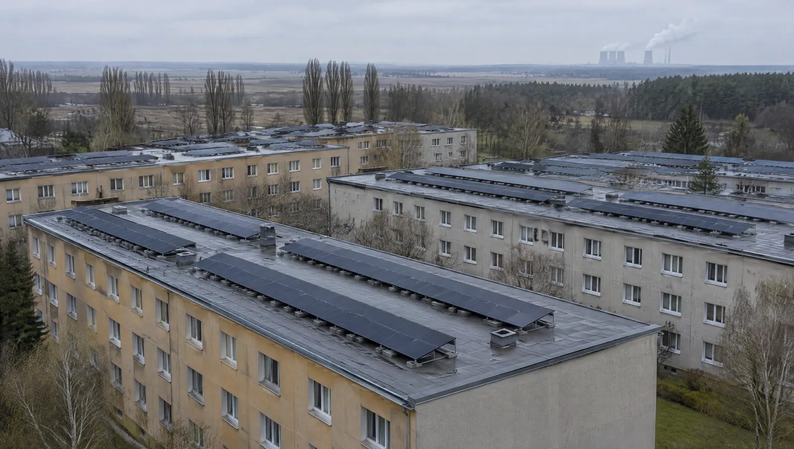 Forst und Guben sind zweimal entstanden: Im Frühjahr 1945 lagen 85 beziehungsweise fast 90 Prozent in Trümmern. Was heute in den Innenstädten steht, ist überwiegend Wiederaufbau und DDR-Wohnungsbau, also Zeilen und Blöcke mit großen, gleichförmigen Dachflächen. Ein Flachdach gibt die Ausrichtung frei, weil die Module aufgeständert werden. Dafür verlangt es eine Aussage zur Statik, zum Zustand der Dachhaut und zur Ballastierung, bevor das erste Gestell gesetzt wird. Blick von oben auf parallele Wohnzeilen im Landkreis Spree-Neiße: breite Flachdächer mit grauer Bitumenbahn, darauf lange Reihen flach aufgeständerter dunkler Module mit Wartungsgassen dazwischen. Dahinter die flache Lausitzer Ebene mit Pappelreihen, Kiefernwald und fernen Dampffahnen unter bedecktem Frühlingshimmel.