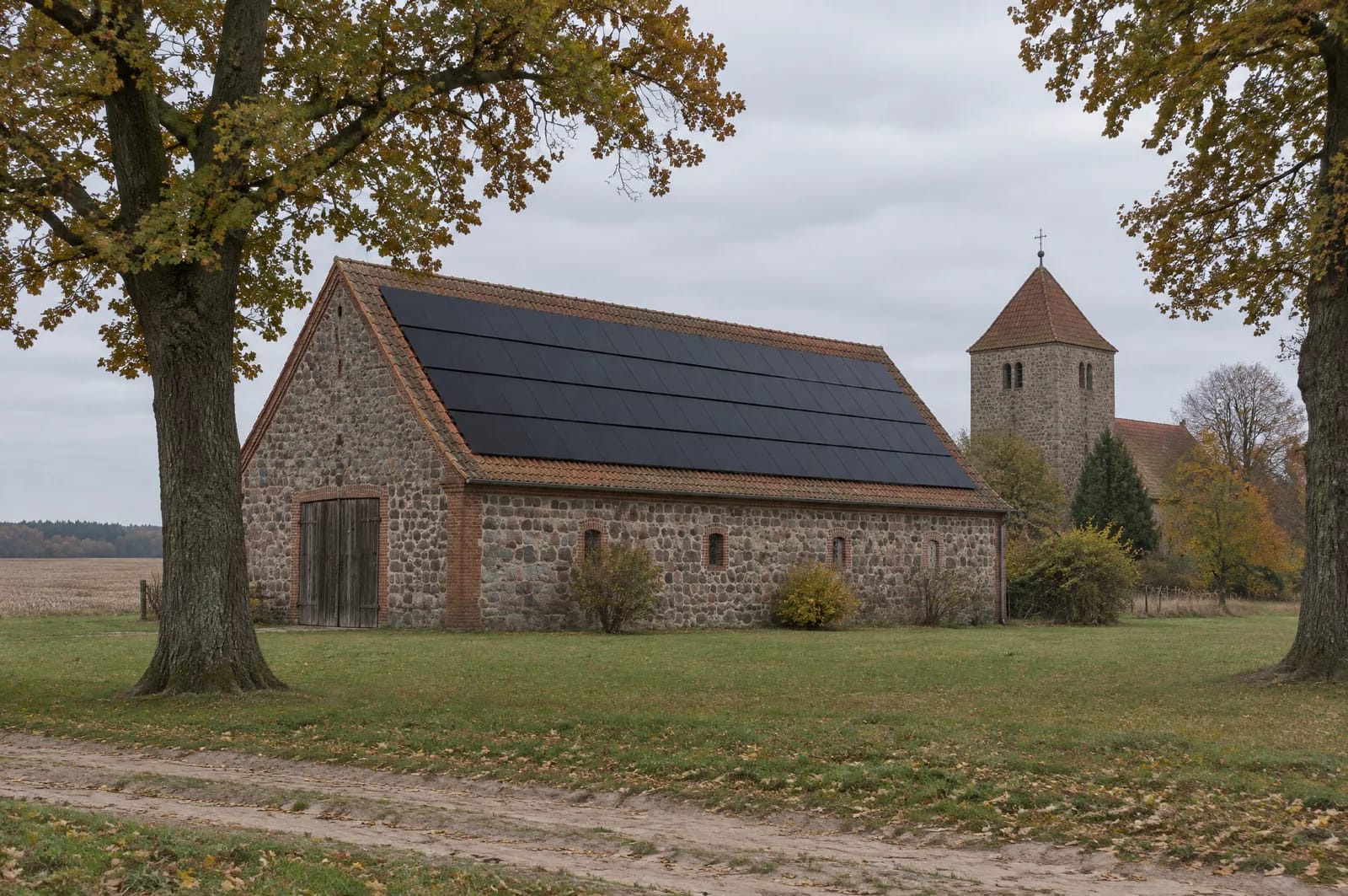 Der Niedere Fläming im Süden des Landkreises Teltow-Fläming ist eine ältere Moränenlandschaft der Saale-Kaltzeit, dünn besiedelt: in Ihlow dreizehn Einwohner je Quadratkilometer, in Dahmetal zehn. Knapp die Hälfte der Kreisfläche wird landwirtschaftlich genutzt, von annähernd vierhundert Betrieben. Entsprechend prägen Hofstellen das Bild — Wohnhaus, Stall und Scheune getrennt um einen Hof. Das Scheunendach ist dort die größte Fläche am Stück und oft die älteste. Ob seine Sparren eine zweite Last aufnehmen, entscheidet sich vor dem Angebot, nicht danach. Nahe Ansicht einer langen Feldsteinscheune im Niederen Fläming, Landkreis Teltow-Fläming: steiles Satteldach mit rotbraunen Biberschwanzziegeln, auf der Südseite ein durchgehendes Feld schwarzer Module in vier Reihen; davor ein Dorfanger mit alten Eichen, dahinter der Feldsteinturm einer Dorfkirche.