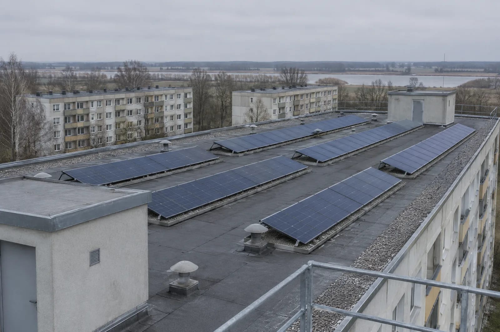 Prenzlau, Schwedt und Gartz haben 1945 ihre Innenstädte verloren. In Prenzlau war bei 716 von 832 Grundstücken der Innenstadt die Bebauung zerstört; der Wiederaufbau begann 1952, hauptsächlich mit Plattenbauten. Schwedt war zu rund 85 Prozent zerstört und hat seit 1989 über vierzig Prozent seiner Einwohner verloren — ganze Straßenzeilen wurden zurückgebaut. Was dort oben liegt, ist Flachdach: freie Ausrichtung, aber eine Aussage zu Statik, Dachhaut und Ballast gehört vor die Modulzahl. Blick von einem Nachbardach auf das Flachdach eines fünfgeschossigen Wohnblocks im Landkreis Uckermark: dunkle Bitumenbahnen, Lüfterhauben und ein Treppenhauskopf, darauf aufgeständerte Modulreihen in Ost-West-Aufstellung auf Ballastwannen, dazwischen Wartungsgassen; im Hintergrund Plattenbauzeilen und kahle Linden.