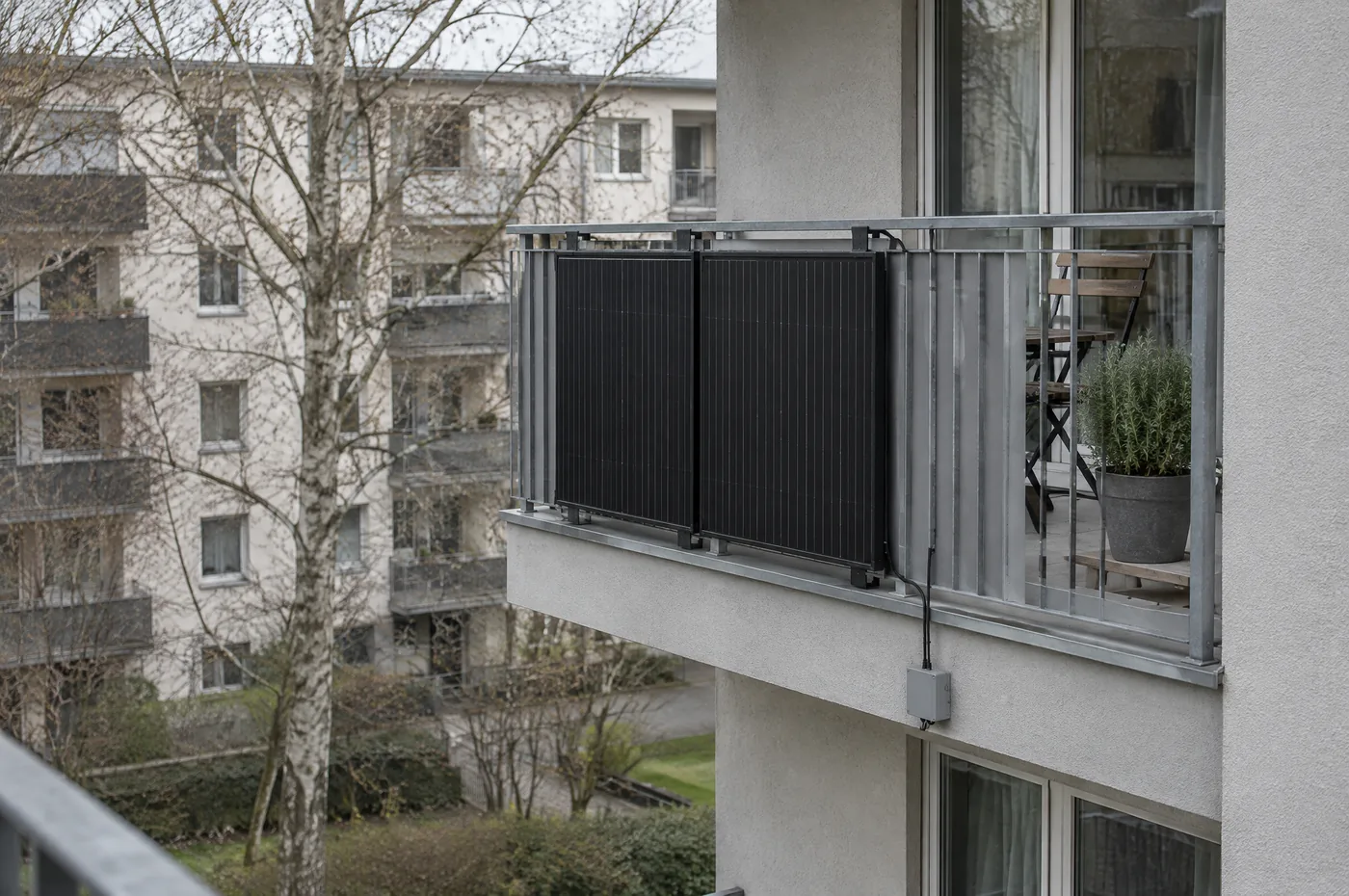 Balkonkraft 2025: Mini Solaranlagen im Fokus Zwei schmale schwarze Solarmodule, flach aussen an die Metallbruestung eines Balkons im Obergeschoss geschraubt; ein schwarzes Kabel fuehrt nach unten in einen kleinen grauen Kasten an der Fassade. Dahinter der Balkon mit Holzstuhl und Kraeutertopf, gegenueber ein Wohnblock und eine kahle Birke.