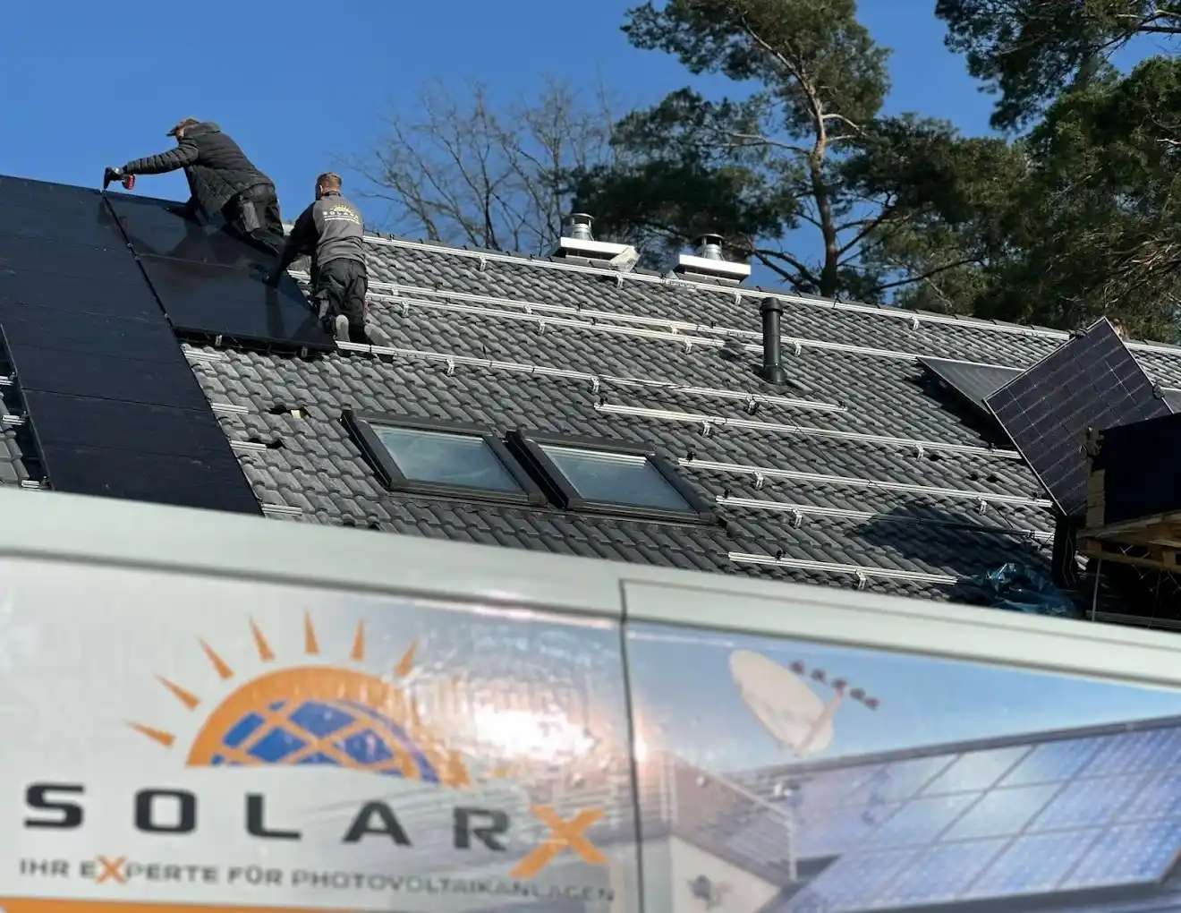 Solaranlage-SolarX – Wir stellen Ihnen die SolarX GmbH vor! Zwei Monteure auf einem grauen Ziegeldach: der vordere schraubt mit dem Akkuschrauber ein schwarzes Modul fest, der zweite kniet im Arbeitsshirt daneben auf den bereits montierten Schienen; im Vordergrund das Dach eines Transporters mit Sonnen-Logo und dem Schriftzug „SolarX — Ihr Experte fuer Photovoltaikanlagen".